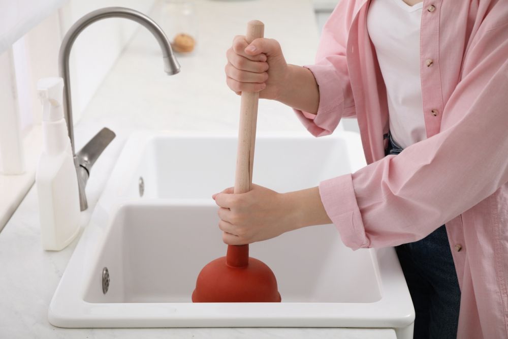 Person using a plunger to unclog a kitchen sink, wearing a pink shirt and standing by a faucet.
