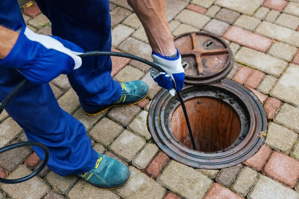Worker cleaning drain with rod on cobblestone pavement, wearing gloves and blue uniform.