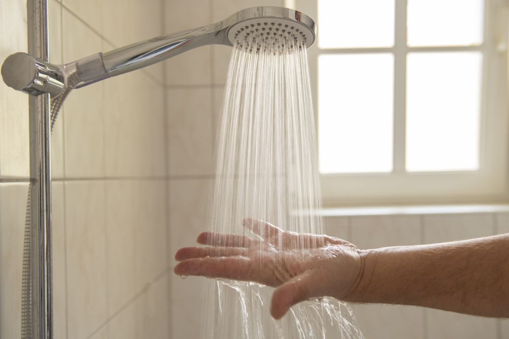 Hand testing water from showerhead in bright bathroom with tiled walls and window in background.