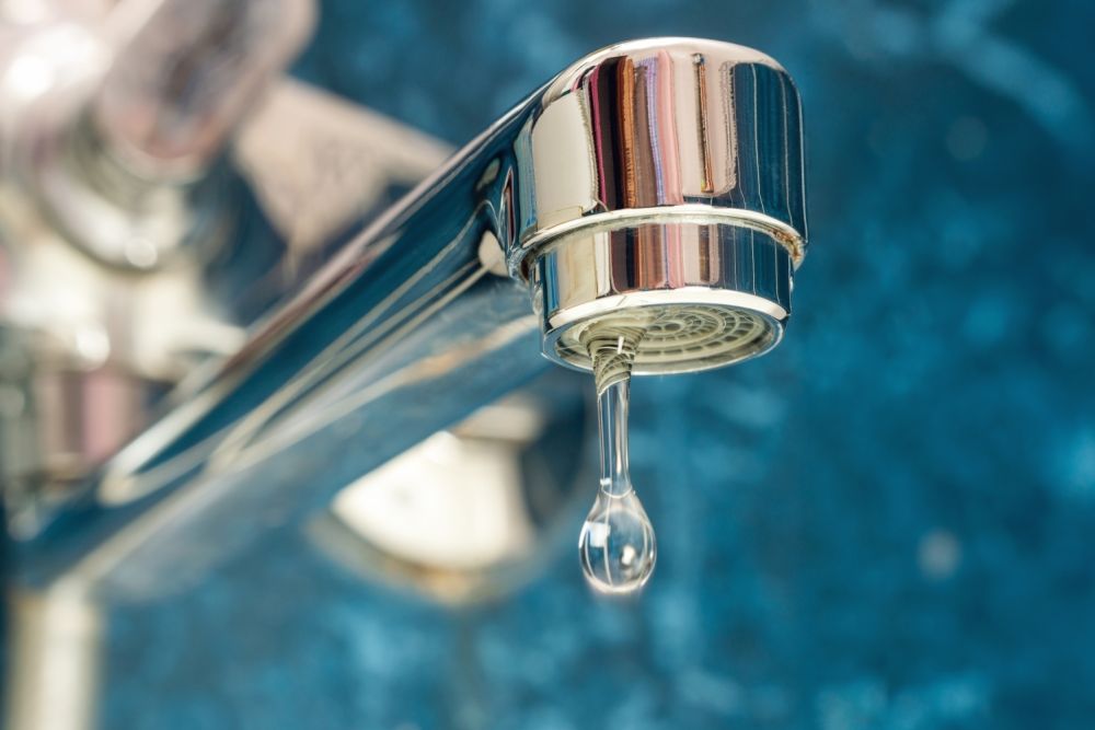 Close-up of a leaking faucet with water droplet, highlighting waste and plumbing issues on a blue background.