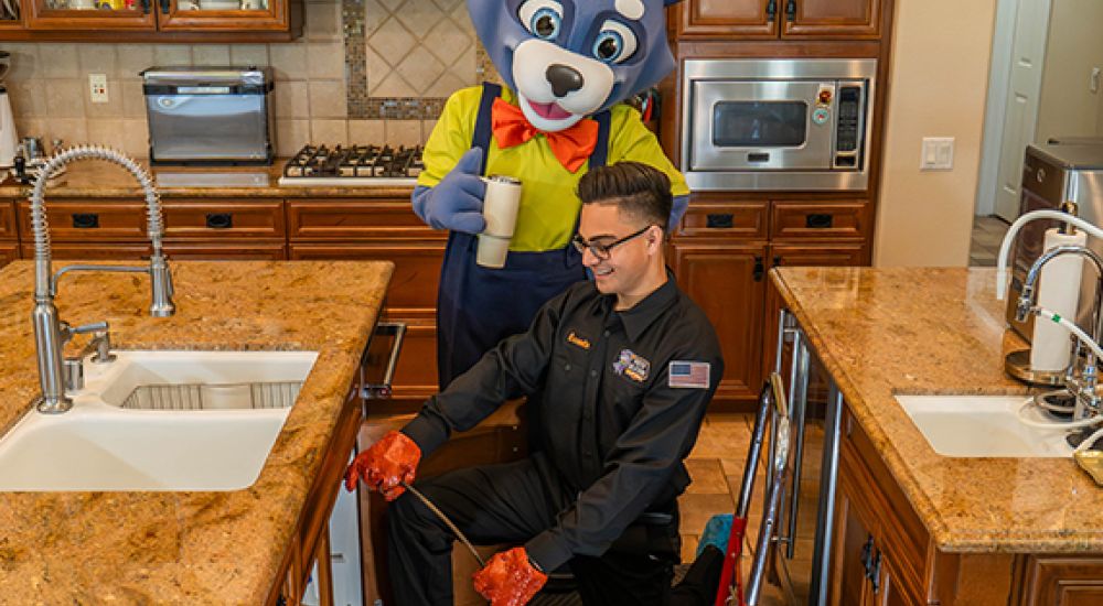 Plumber fixing kitchen sink as mascot watches with a drink, showcasing fun and efficient service.