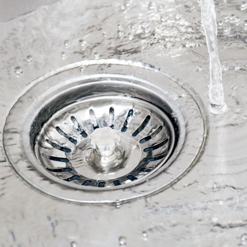 Water flowing down a stainless steel sink drain, creating ripples and bubbles on the surface.